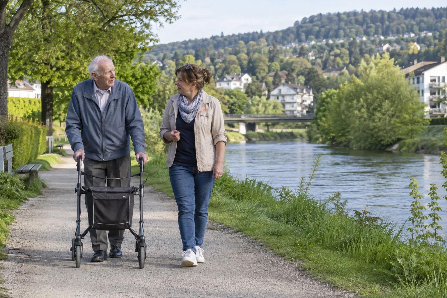 Ein älterer Mann mit Rollator spaziert mit einer Begleitperson an einem Fluss